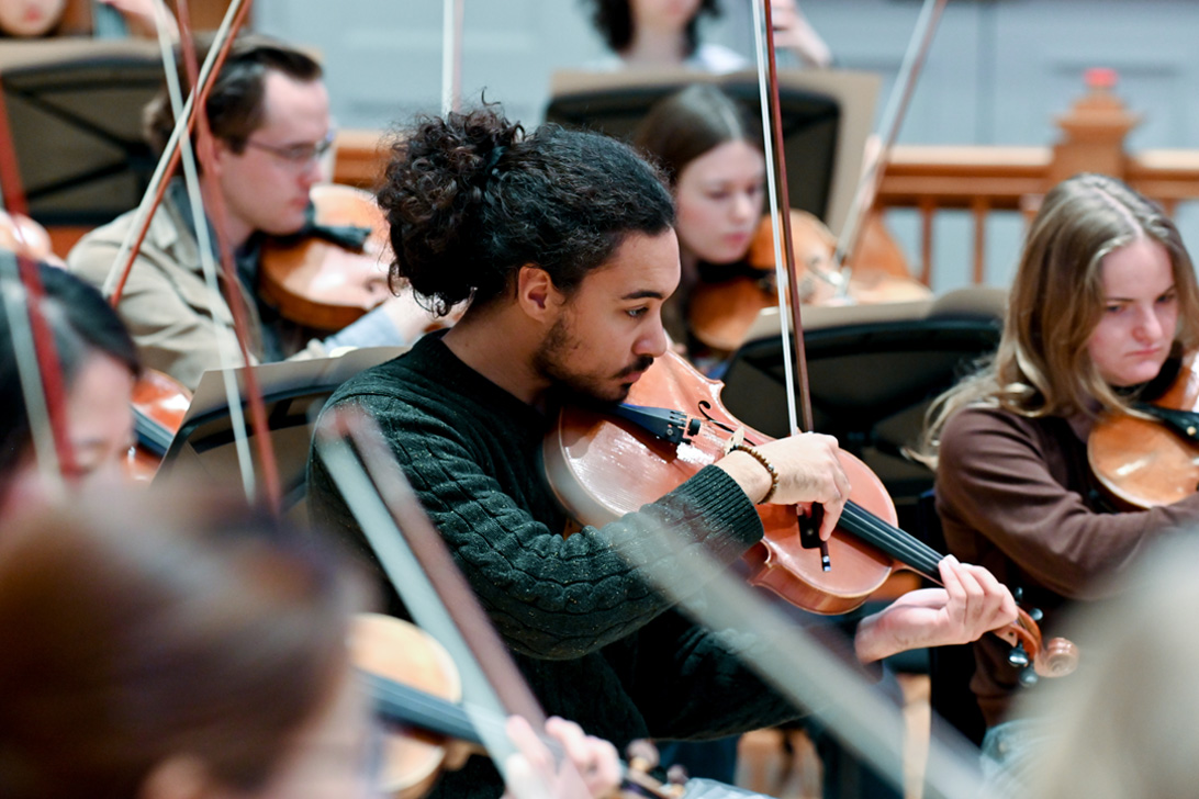 A male student, wearing smart attire, playing the viola in a string section in an orchestra performance.